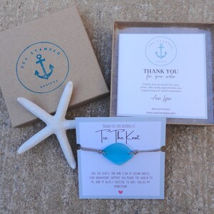 Puede incluir: Un brazalete de vidrio de mar azul con un cord&oacute;n marr&oacute;n y el texto "Tie The Knot" en una tarjeta blanca. La tarjeta tambi&eacute;n incluye un mensaje sobre el suave flujo y reflujo de las olas del oc&eacute;ano.