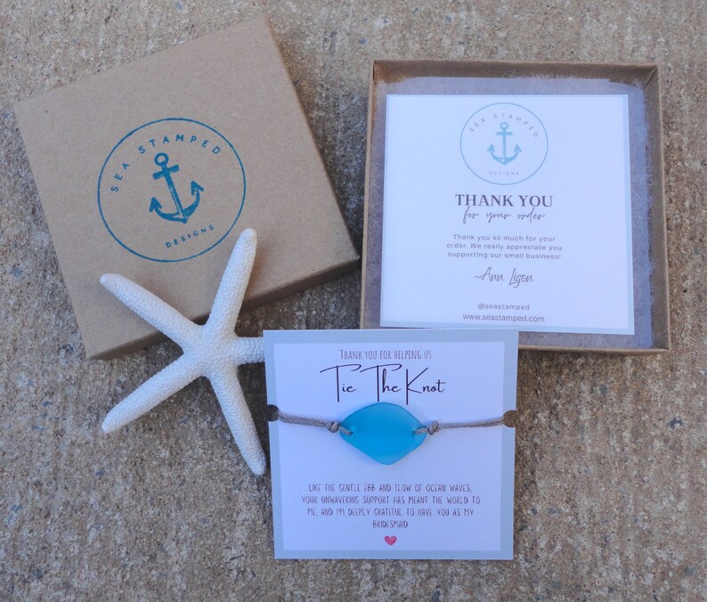 Puede incluir: Un brazalete de vidrio de mar azul con un cord&oacute;n marr&oacute;n y el texto "Tie The Knot" en una tarjeta blanca. La tarjeta tambi&eacute;n incluye un mensaje sobre el suave flujo y reflujo de las olas del oc&eacute;ano.