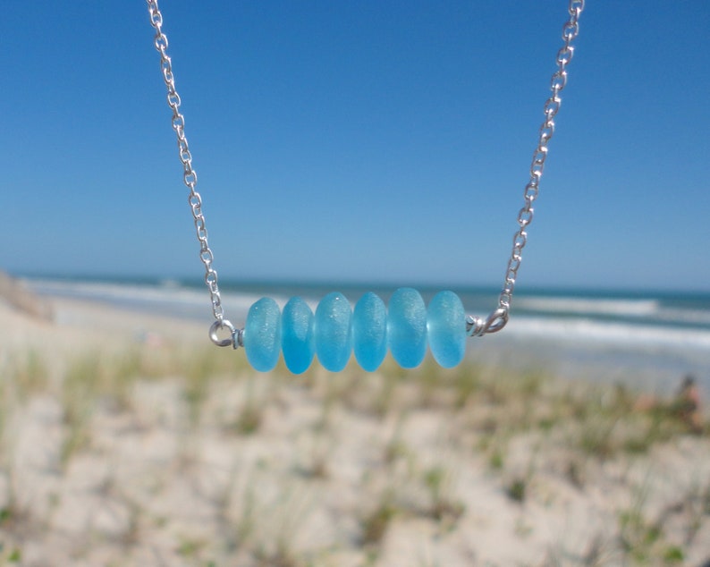 May include: A silver chain necklace with a row of six light blue sea glass beads. The necklace is photographed against a blurred background of a beach.