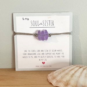 Może przedstawiać: Bransoletka z fioletowego szkła morskiego z sznurkiem w kolorze szarym i kartką z napisem "To my Soul Sister" oraz wiadomością o przypływie i odpływie fal oceanu. Na kartce znajduje się r&oacute;wnież adres strony internetowej www.seastamped.com.
