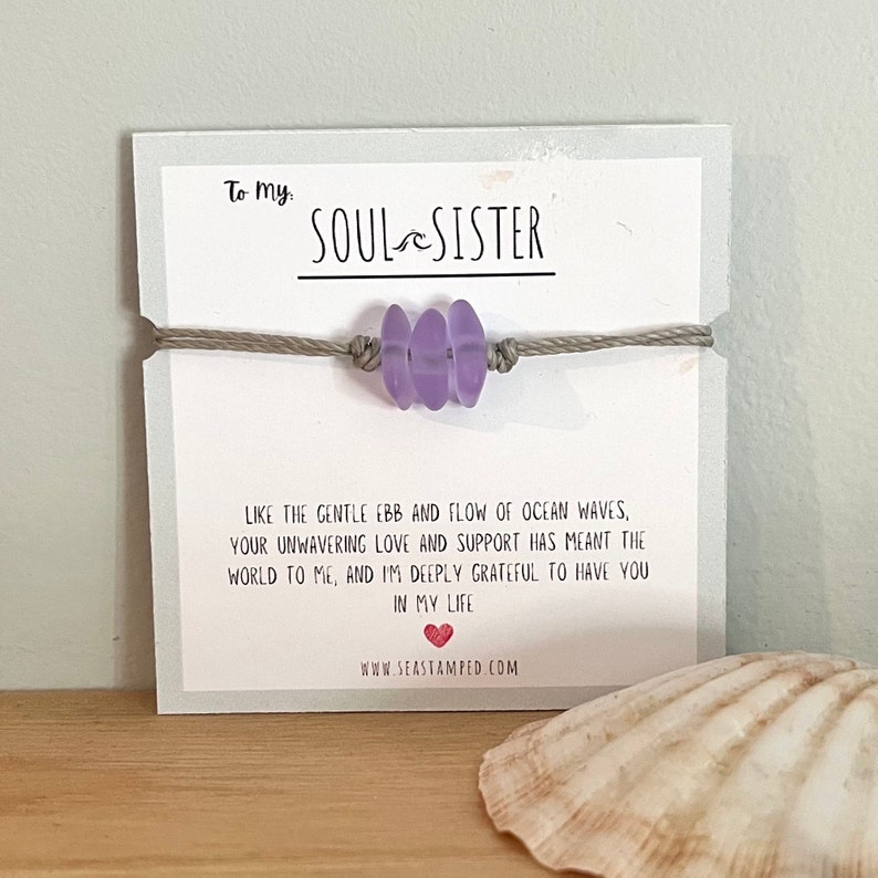 Może przedstawiać: Bransoletka z fioletowego szkła morskiego z sznurkiem w kolorze szarym i kartką z napisem "To my Soul Sister" oraz wiadomością o przypływie i odpływie fal oceanu. Na kartce znajduje się r&oacute;wnież adres strony internetowej www.seastamped.com.