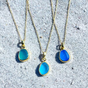 May include: Three gold necklaces with different shades of blue sea glass pendants. The necklaces are on a white sandy surface.