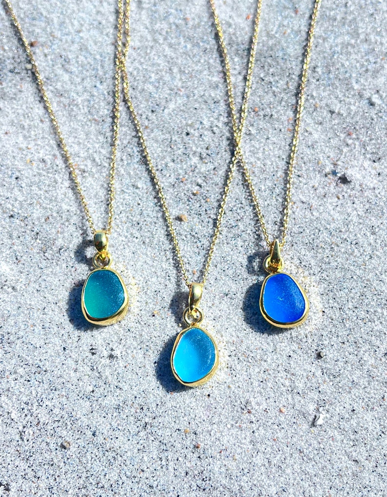 May include: Three gold necklaces with different shades of blue sea glass pendants. The necklaces are on a white sandy surface.