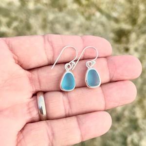 May include: A pair of teardrop-shaped earrings with a light blue, sea glass appearance. The earrings have a silver-colored metal border and hook. The background is a blurred, natural outdoor setting.