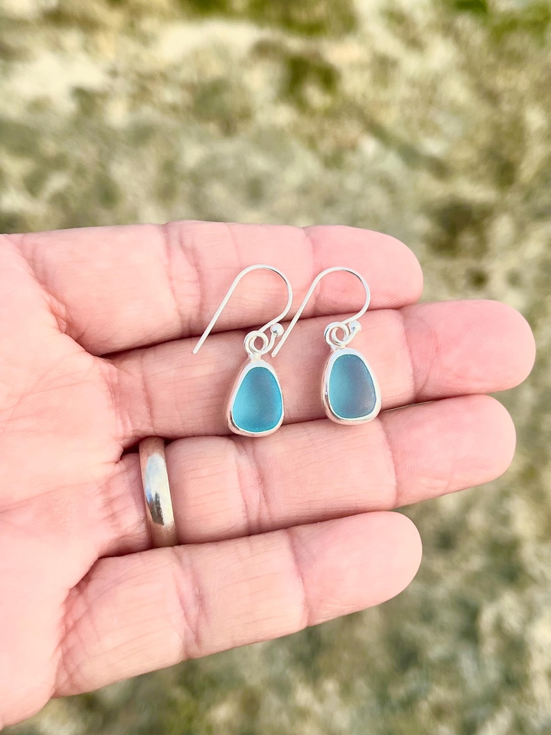 May include: A pair of teardrop-shaped earrings with a light blue, sea glass appearance. The earrings have a silver-colored metal border and hook. The background is a blurred, natural outdoor setting.