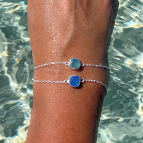 May include: Two silver chain bracelets, each featuring a single small, smooth, rounded stone. One stone is a pale, translucent aqua blue, while the other is a vibrant, solid cobalt blue. The bracelets are delicate, with thin metal links.