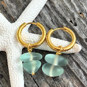 Può includere: Orecchini a cerchio color oro con pendenti in vetro marino. Ogni orecchino presenta un cerchio dorato con due pezzi di vetro marino ovali azzurri appesi. Gli orecchini sono esposti su una stella marina bianca e una superficie di legno invecchiato.