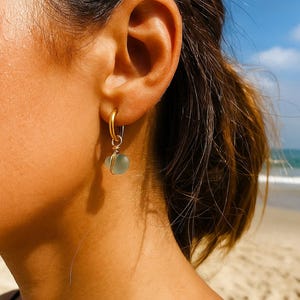 May include: A gold hoop earring with a small, light blue, sea glass pendant. The earring is attached to an ear, with the beach and ocean visible in the background. The earring is a simple, elegant design.