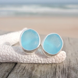 May include: A pair of light blue sea glass stud earrings with a silver bezel setting. The earrings are displayed on a white starfish against a blurred background of the ocean and sky. The earrings are a unique jewelry item.
