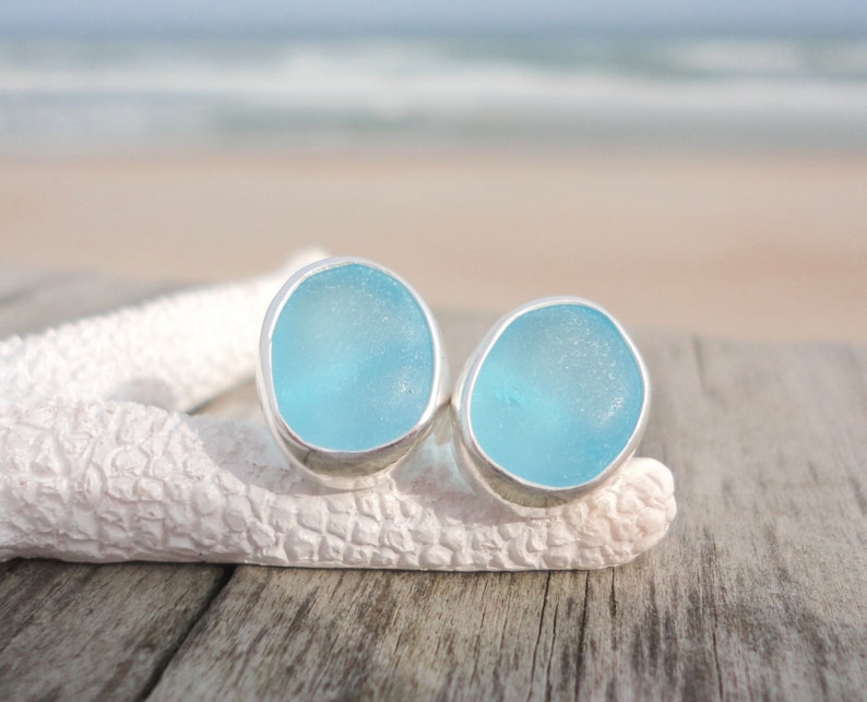 May include: A pair of light blue sea glass stud earrings with a silver bezel setting. The earrings are displayed on a white starfish against a blurred background of the ocean and sky. The earrings are a unique jewelry item.