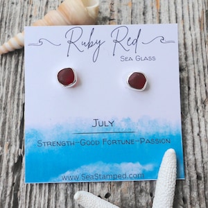 May include: A pair of ruby red sea glass stud earrings with silver-colored settings. The earrings are displayed on a white card with the text "Ruby Red Sea Glass" and "July: Strength-Good Fortune-Passion". A seashell and starfish are also in the image.