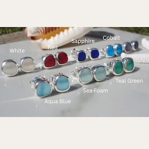 May include: A collection of colorful stud earrings in various shades, including white, red, sapphire, cobalt, teal green, sea foam, and aqua blue. Each earring features a square-shaped, polished stone set in a silver metal frame. The earrings are displayed on a reflective surface.