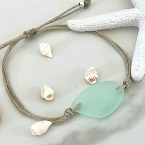 Peut inclure: Un bracelet en cordon marron avec un charme en verre de mer vert clair et des coquillages blancs.