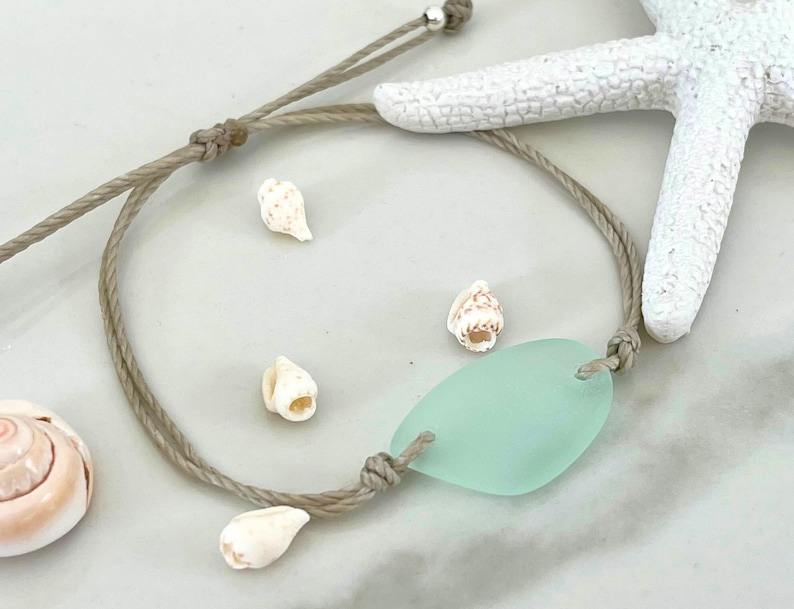 Peut inclure: Un bracelet en cordon marron avec un charme en verre de mer vert clair et des coquillages blancs.