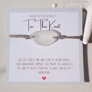Puede incluir: Una tarjeta blanca con el texto "Tie The Knot" y un mensaje agradeciendo a alguien por ayudarlos a atar el nudo. La tarjeta tiene un amuleto de vidrio de mar atado a una cuerda beige.