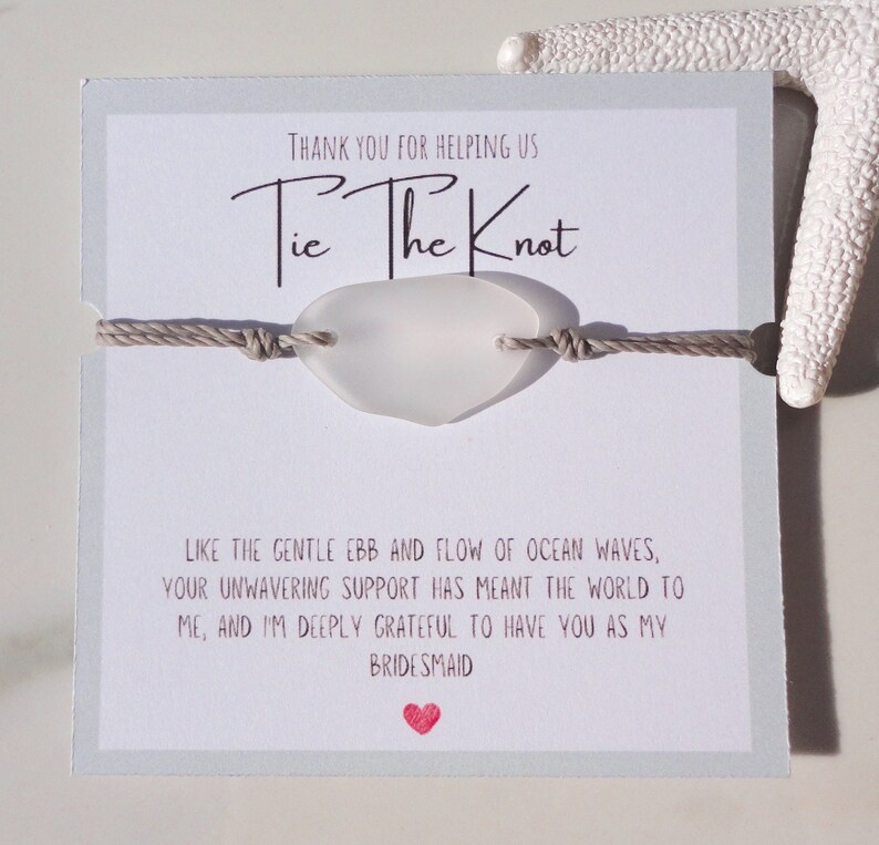 Puede incluir: Una tarjeta blanca con el texto "Tie The Knot" y un mensaje agradeciendo a alguien por ayudarlos a atar el nudo. La tarjeta tiene un amuleto de vidrio de mar atado a una cuerda beige.