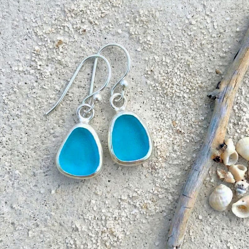 May include: A pair of silver-toned drop earrings with a teardrop shape. Each earring features a vibrant turquoise-blue glass insert. The earrings are displayed on a sandy surface, suggesting a beach theme.