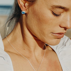 Op de afbeelding: Een zilveren ketting met een blauwe, traanvormige hanger. De hanger is gemaakt van een gladde, gepolijste steen. De ketting wordt gedragen door een persoon met donker haar.