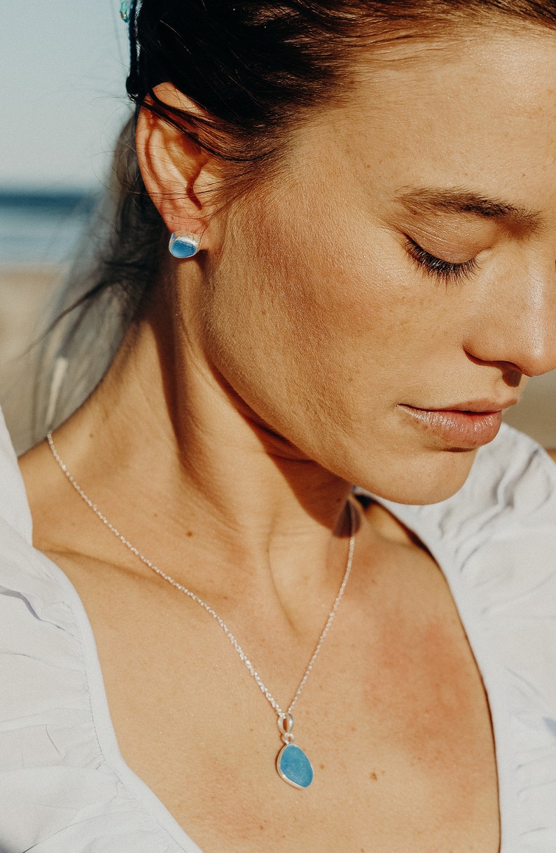 Op de afbeelding: Een zilveren ketting met een blauwe, traanvormige hanger. De hanger is gemaakt van een gladde, gepolijste steen. De ketting wordt gedragen door een persoon met donker haar.