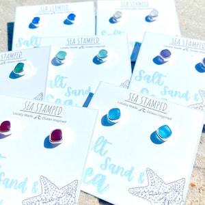 May include: A collection of colorful stud earrings on white cards, each with a silver setting. The earrings come in various colors, including blue, green, purple, and red. The cards are printed with the text "SEA STAMPED" and "Locally Made Ocean Inspired".