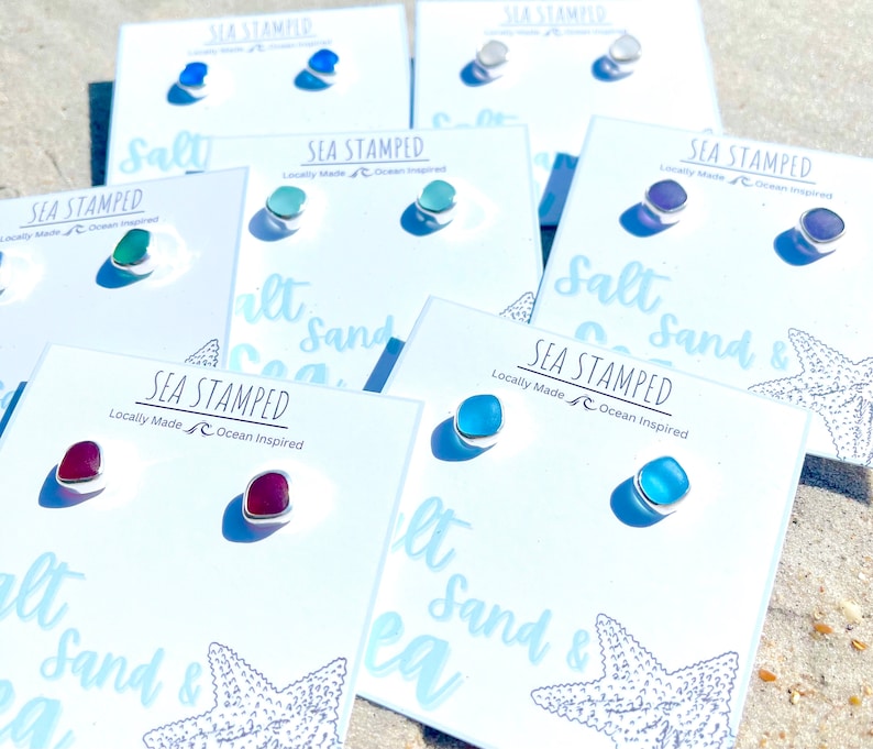 May include: A collection of colorful stud earrings on white cards, each with a silver setting. The earrings come in various colors, including blue, green, purple, and red. The cards are printed with the text "SEA STAMPED" and "Locally Made Ocean Inspired".