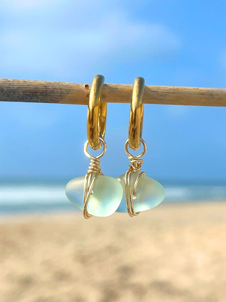 May include: A pair of gold hoop earrings with sea glass pendants. The sea glass is a pale blue-green color, and the hoops are a shiny gold. The earrings are hanging on a wooden stick, with a beach and ocean background.