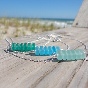 May include: Two silver necklaces with a row of sea glass beads. The top necklace has green sea glass beads and the bottom necklace has blue sea glass beads.
