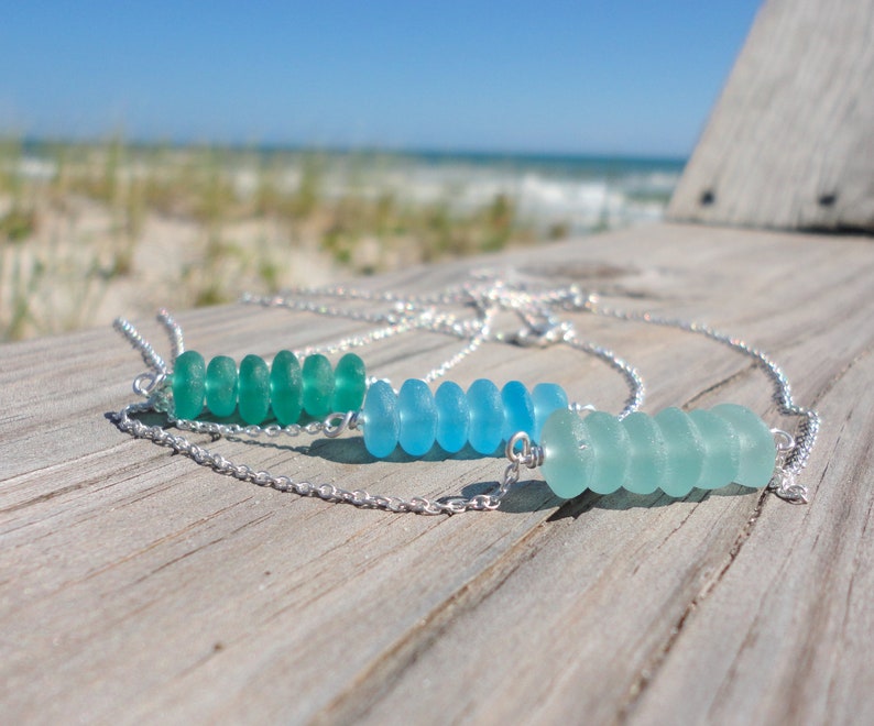 May include: Two silver necklaces with a row of sea glass beads. The top necklace has green sea glass beads and the bottom necklace has blue sea glass beads.