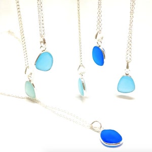 Op de afbeelding: Vijf zilveren kettingen met blauwe zeeglas hangers. De hangers zijn in verschillende tinten blauw, van licht tot donker.