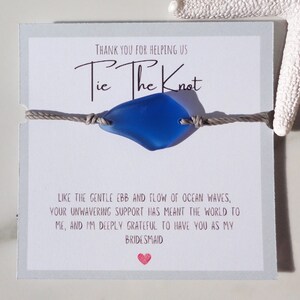 Puede incluir: Una tarjeta gris con el texto "Tie The Knot" y un dije de vidrio de mar azul en un cord&oacute;n gris. La tarjeta tambi&eacute;n incluye el texto "Gracias por ayudarnos" y "Como el suave vaiv&eacute;n de las olas del oc&eacute;ano, tu apoyo inquebrantable ha significado el mundo para m&iacute;, y estoy profundamente agradecida de tenerte como mi dama de honor."