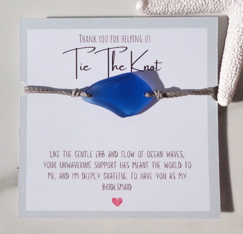 Puede incluir: Una tarjeta gris con el texto "Tie The Knot" y un dije de vidrio de mar azul en un cord&oacute;n gris. La tarjeta tambi&eacute;n incluye el texto "Gracias por ayudarnos" y "Como el suave vaiv&eacute;n de las olas del oc&eacute;ano, tu apoyo inquebrantable ha significado el mundo para m&iacute;, y estoy profundamente agradecida de tenerte como mi dama de honor."