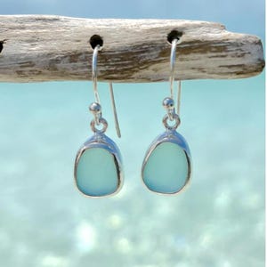 May include: A pair of silver dangle earrings with light blue, sea glass-like stones. The earrings are teardrop-shaped and hang from silver hooks. The background is a blurred image of the ocean and sky.