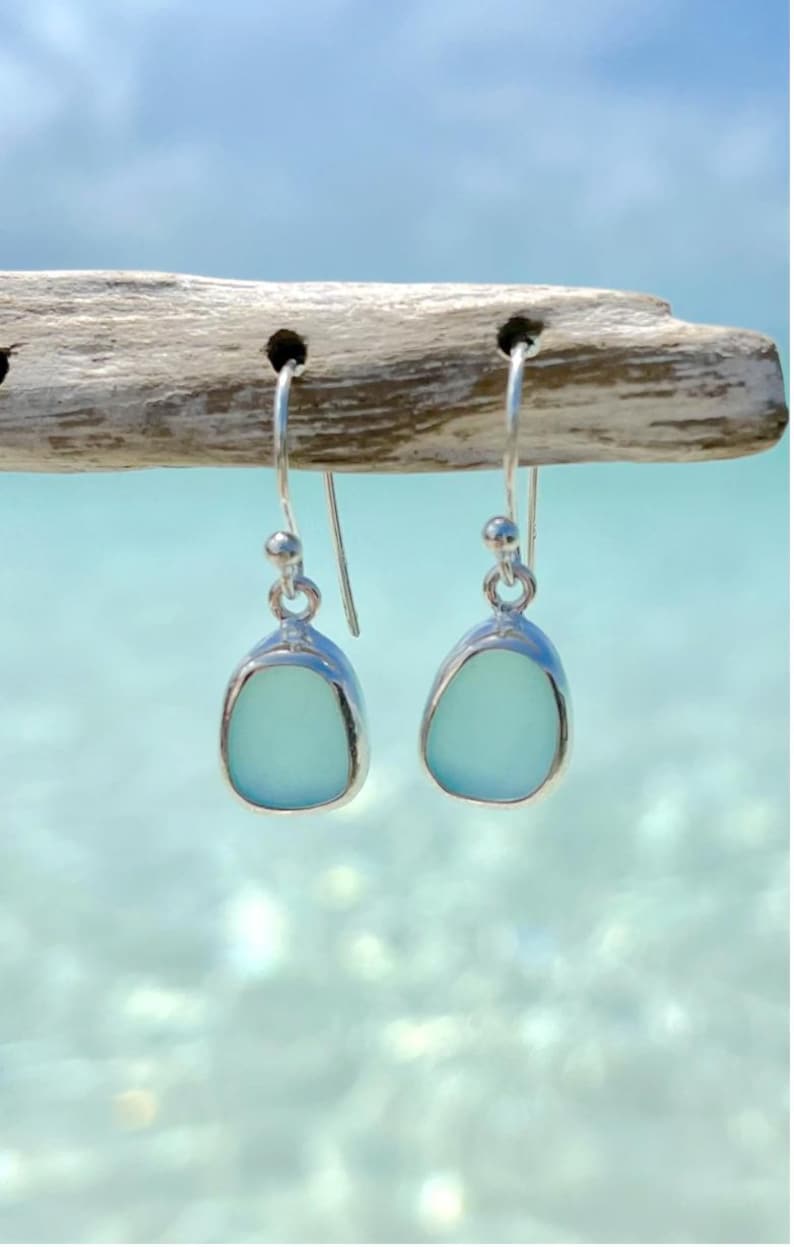 May include: A pair of silver dangle earrings with light blue, sea glass-like stones. The earrings are teardrop-shaped and hang from silver hooks. The background is a blurred image of the ocean and sky.