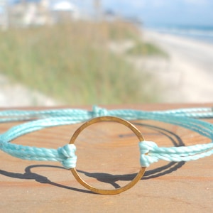 May include: A light blue braided cord bracelet with a gold circle charm. The bracelet is on a wooden surface.