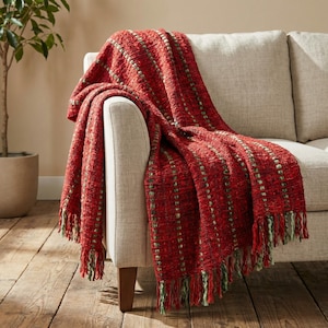 May include: A red woven throw blanket with green accents and fringe is draped over a beige sofa. The blanket has a textured appearance with vertical stripes of green dots. The fringe is a mix of red and green.