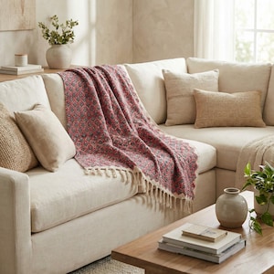 May include: A patterned red and grey throw blanket with a fringe is draped over a cream-coloured sectional sofa. Beige throw cushions and a wooden coffee table with books and a vase complete the cosy living room scene.