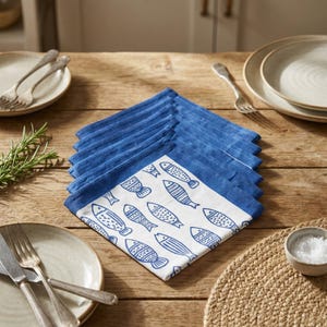 Puede incluir: Un conjunto de servilletas de tela azules y blancas con un diseño de peces, dispuestas sobre una mesa de madera. Las servilletas tienen un borde azul y un centro blanco adornado con ilustraciones de peces azules. El conjunto se completa con platos, cubiertos y un pequeño cuenco de sal.
