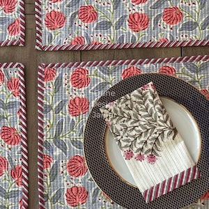 Può includere: Set di tovagliette floreali con set di tovaglioli abbinati. Le tovagliette presentano un motivo ripetuto di fiori rossi e foglie blu su uno sfondo trapuntato. Un piatto e un tovagliolo sono appoggiati su una delle tovagliette.