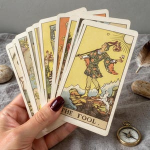 Classic Rider Waite tarot cards held in hand featuring The Fool card, vintage tarot reading setup with compass and natural fabric background