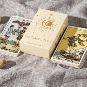 Classic Rider Waite tarot deck with The Fool card and Two of Cups card beside premium tarot box on fabric surface