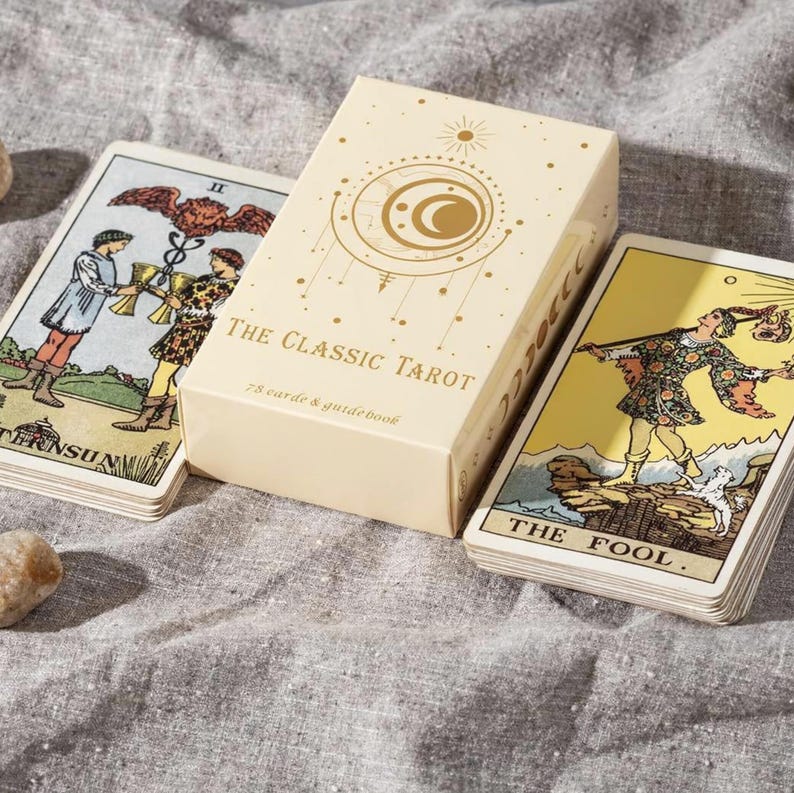 Classic Rider Waite tarot deck with The Fool card and Two of Cups card beside premium tarot box on fabric surface