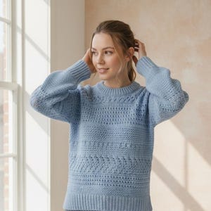 May include: A light blue sweater with a textured knit design. The sweater features a ribbed hem and cuffs, and a crew neck. The knit pattern includes horizontal and diamond-shaped sections. The sleeves are long and the sweater is made of a soft material.