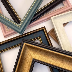 Könnte beinhalten: Eine Sammlung von Bilderrahmen in verschiedenen Farben, darunter Türkis, Schwarz, Gold und Rosa. Die Rahmen haben unterschiedliche Stile, einige mit aufwendigen Golddetails. Die Rahmen sind auf einer weißen Oberfläche angeordnet.