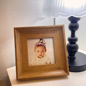 May include: A square wooden photo frame displaying a portrait of a baby girl wearing a pink headband. The frame is made of light brown wood and is on a white surface next to a black lamp.