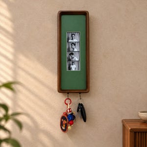 Può includere: Un portachiavi da parete rettangolare in legno con cornice in legno scuro e pannello interno verde. Il centro mostra una striscia verticale di quattro fotografie ritratto in bianco e nero. Due ganci in metallo sostengono mazzi di chiavi.