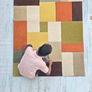May include: A rectangular area rug featuring a geometric pattern of solid color blocks in shades of olive green, mustard yellow, burnt orange, cream, and dark brown. The rug has a textured, tufted surface with clean, straight edges and seams.
