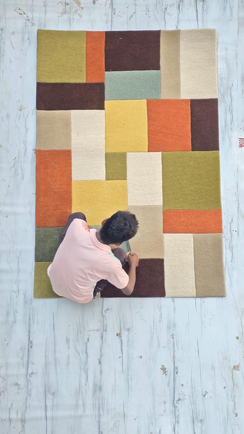 May include: A rectangular area rug featuring a geometric pattern of solid color blocks in shades of olive green, mustard yellow, burnt orange, cream, and dark brown. The rug has a textured, tufted surface with clean, straight edges and seams.