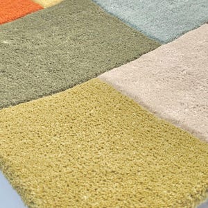 May include: A tufted rug featuring a grid of square sections in various colors including mustard yellow, olive green, light blue, orange, and beige. The rug has a thick, textured pile surface with visible fibers and a distinct block pattern design.