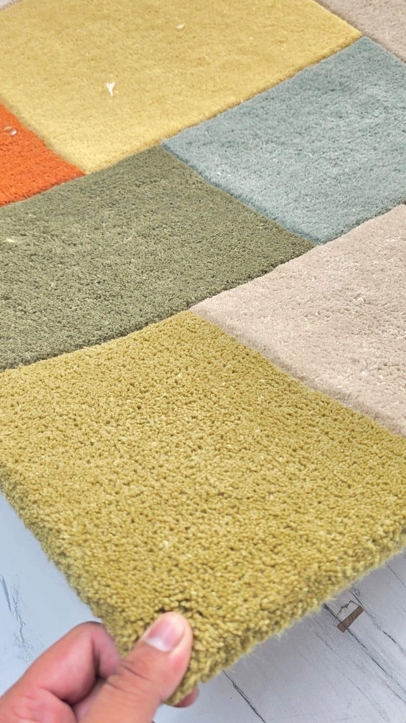 May include: A tufted rug featuring a grid of square sections in various colors including mustard yellow, olive green, light blue, orange, and beige. The rug has a thick, textured pile surface with visible fibers and a distinct block pattern design.