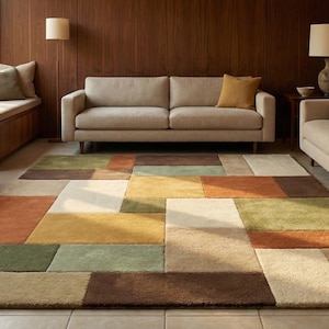 Op de afbeelding: Een woonkamer met een beige bank, een patroonkleed en een houten salontafel. Het kleed heeft een geometrisch ontwerp met bruine, groene, oranje en crèmekleurige vierkanten. De kamer heeft grote ramen en houten lambrisering.